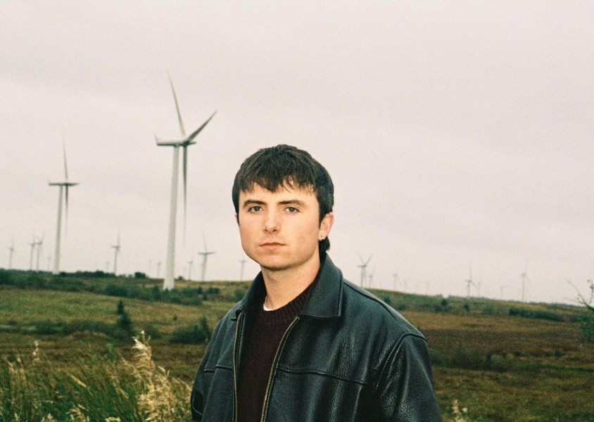 A guy in a brown jumper and black leather jacket stood in a field in front of wind turbinesx